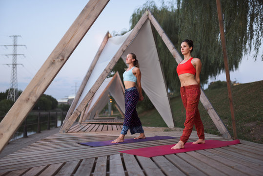 Two female friends practicing yoga on the river shore
