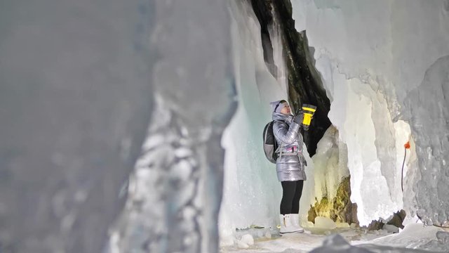 Travel woman on ice cave of Lake Baikal. Trip to winter island. Girl backpacker is walking of ice grot. Traveler looks at beautiful ice grotto. Hiker wears sports glasses, silver jacket, backpack, led