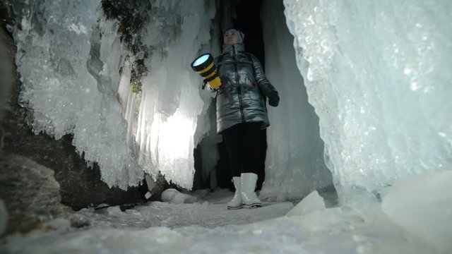 Travel woman on ice cave of Lake Baikal. Trip to winter island. Girl backpacker is walking of ice grot. Traveler looks at beautiful ice grotto. Hiker wears sports glasses, silver jacket, backpack, led