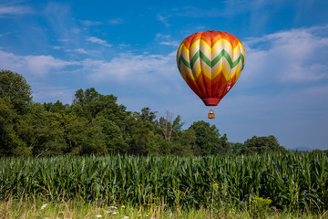 Red, Yellow, Orange, Green, and White Teardrop Shaped Hot Air Balloon Over Corn Field on Sunny Day with Trees and Cloudy Blue Sky in Background