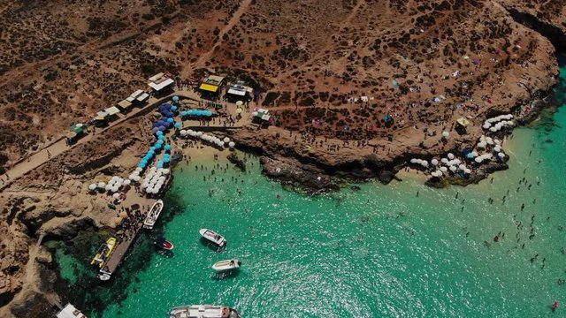 Flying over a busy beach at the Blue Lagoon on Comino Island, Malta. This aerial shot emphasises the clear water, and gives the video a tropical touch. Lots of boats, swimmers and colourful parasols.