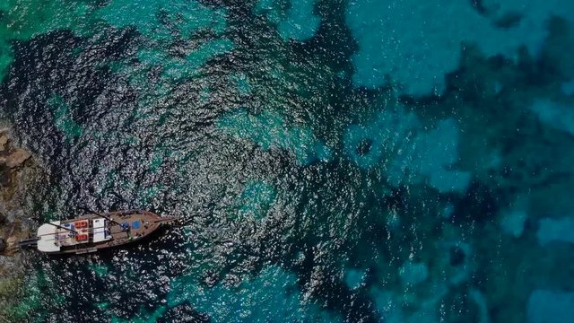 A beautiful overhead shot of a sailing ship next to a rocky shore. The overhead emphasises the sea with its blue and light blue pattern, therefore giving the shot a tropical feel.