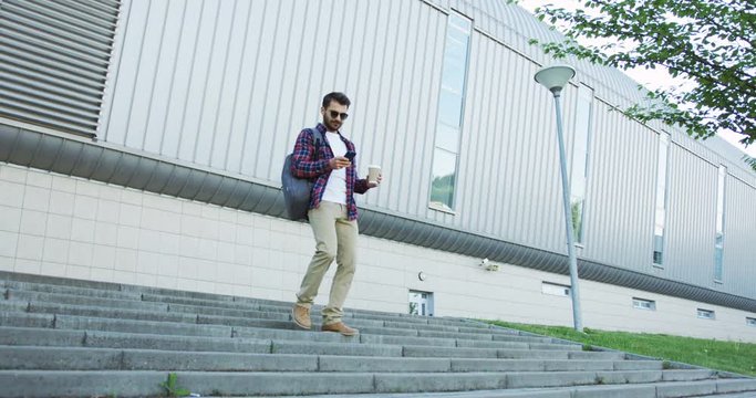 Attractive smiled Caucasian male student walking the street stairs down with a coffee in a hand and scrolling on his smartphone.
