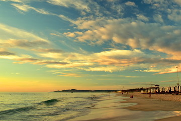 Naklejka premium Sandy beach on the background of a beautiful cloudy sky with golden light. Perspective of the coast. People came to meet the dawn. Beach umbrellas with deckchairs. Waves on the shore