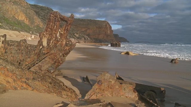 Wide shot of a sunny, sandy beach in the afternoon sunlight covered with corroded pieces of a ship wreck