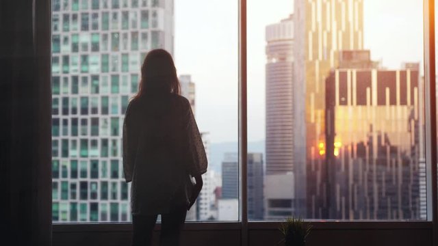 Young pensive woman stands by the big window looking out her apartment on the city buildings during sunset. slow motion. 3840x2160.