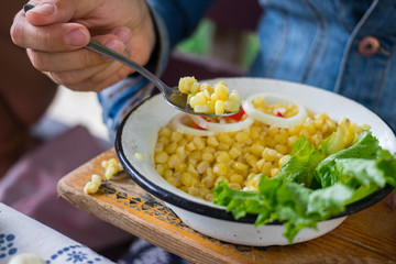 Woman hands holds sweet white corn or yellow corn cooked. Tasting grains cut into metal bowl. Vegetarian lunch. Vegan dinner. Healthy food. Served with green salad leaves.