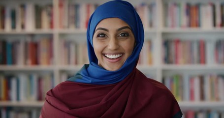 portrait excited young muslim woman smiling enjoying successful education accomplishment wearing hijab in library bookshelf background slow motion