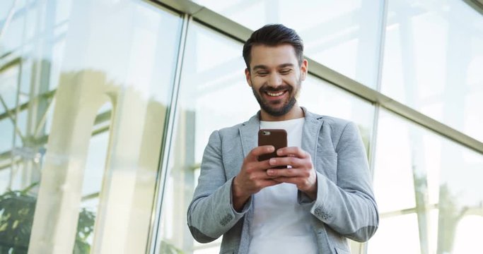 Cheerful young businessman in the official style texting on the smartphone and laughing ner office building.