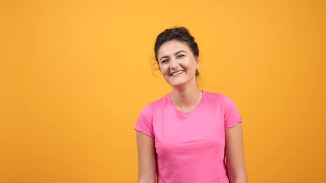 Young woman in pink t-shirt smiling and laughing hard with natural emotions on yellow orange background. Happy carefree attractive girl posing in studio. Slow motion footage