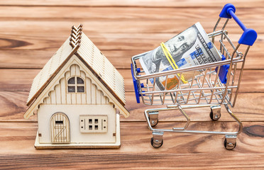 Shopping cart with rolled up dollar bills and model of house on the wooden background.