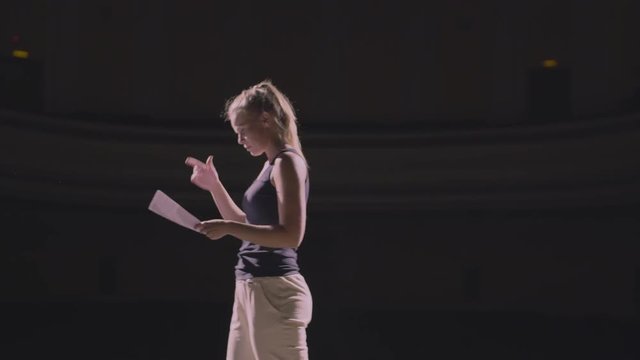 Adult girl walking on the theatre stage and do rehearsal before the premiere. At background empty hall with many rows of seats. The actress concentrated on the script and ready her self for acting