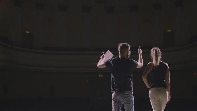 Rehearsal at theatre. Man explain to student actress how to play on the stage on the new performance for public. Dark hall at background. Girl carefully listen the director and say something by answer