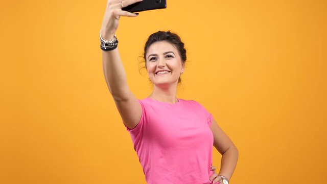 Happy smiling woman in pink t-shirt taking a selfie on yellow orange background