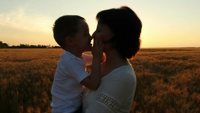 A happy mother holds a child in her arms in a field of wheat against a sunset background. A happy child kisses the mother's face. The concept of a happy family
