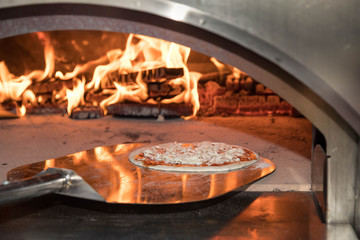 The working furnace for preparation of pizza. Inside burns firewood and big fire rises, on a shovel in the furnace put cheese pizza. Reflections of a flame on a surface