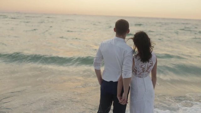 Rear romantic view of the beautiful cou ple of newlyweds softly holding hands and enjoying the sea in the sunset.