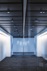 A corridor of business office with glass reflection surface in the middle of the frame with soundproof panels with lamps on the top, white backlit walls, and the grey carpet on the floor