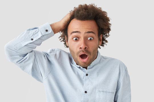 Shocked curly male looks with terrified expression, keeps hand on head, stares with bugged eyes, hears something terrible, dressed in white shirt, poses indoor. Puzzled African American guy.
