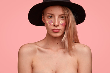 Cropped image of good looking Caucasian female with sparkles on cheeks, has bare shoulders, looks seriously at camera, poses for fashion magazine, isolated over pink background, wears only hat