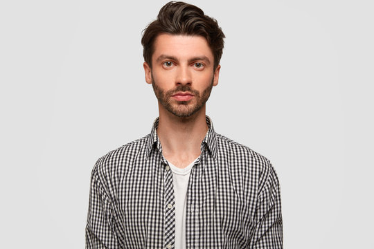 Photo of attractive man with stylish hairdo, has stubble, looks directly at camera seriously, wears checkered shirt, isolated over white studio wall. Confident male manager works, models indoor