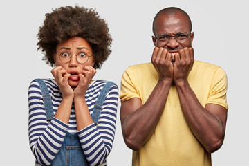 Nervous dark skinned woman and man look anxiously, keep hands near mouthes, notice something...