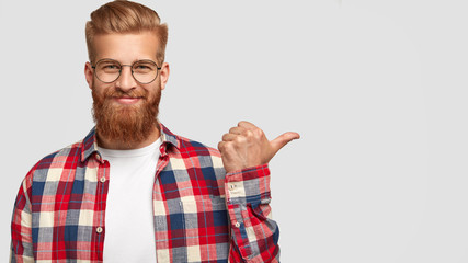 Cheerful bearded male has charming smile, points aside, shows nice place to visit, attracts your...