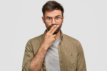 Photo of serious bearded young guy keeps two fingers on mouth, tries to make decision, looks with strict expression, going to punish someone, wears casual clothes, isolated over white background