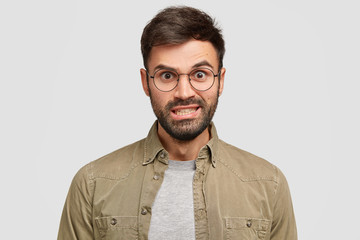 Annoyed Caucasian male raises eyebrows, clenches teeth and looks with anger, wears round spectacles and shirt, expresses negativity, stands against white background. People and emotions concept