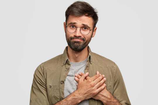 Oh, thank you very much! Attractive unshaven young male keeps hands on heart, expresses gratitude, stands against white studio wall. Friendly bearded Caucasian guy being thankful to best friend