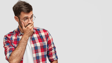 Surprised handsome young male covers mouth with palm, looks with shocked expression aside, notices something strange, dressed in checkered shirt, isolated over white background. Reaction concept