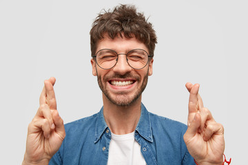 Happy unshaven male with broad smile, shows white teeth, crosses fingers for good luck, being in...