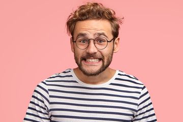 Joyful Caucasian male clenches teeth and looks positively at camera, has curly hair, wears spectacles and striped sweater, isolated over pink background. Happy man freelancer rejoices success