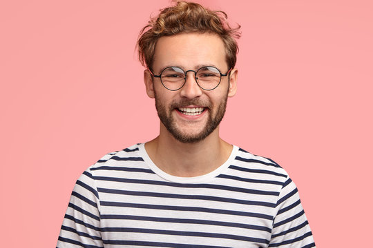 Positive young Caucasian male with pleasant friendly smile, shows white teeth, rejoices new stage in life, wears casual striped sweater and round spectacles, stands alone against pink background.