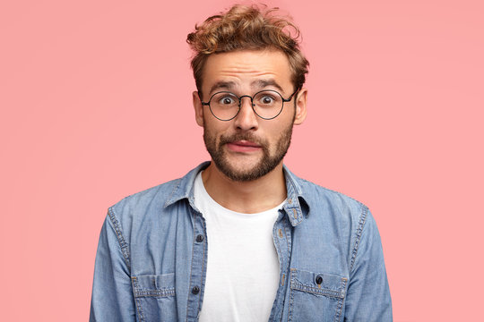 Thrilled curly hipster with puzzled expression purses lips and looks with stunned expression, wears denim shirt, has stubble, isolated over pink background. People, reaction and lifestyle concept