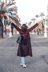 Back view hipster girl with hands up,wearing long bored jacket, bag and earmuffs on city landscape, mock up. Traveler on urban background. Hand raised up. Tourist in Barcelona,  relax lifestyle