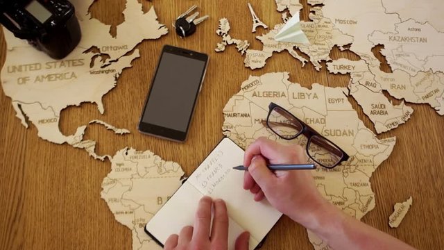 The man is planning a tourist trip to Morocco. Wooden map of the world map on the table, paper airplane on the map, notebook, list of countries