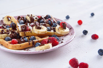 Tasty belgian waffles with blueberries, raspberries, bananas and sugar powder are filled with liquid chocolate.