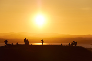 Human silhouettes standing on a hill in front of an orange sunset.