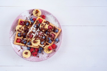 Tasty belgian waffles with blueberries, raspberries, bananas and sugar powder are filled with liquid chocolate. Top view.