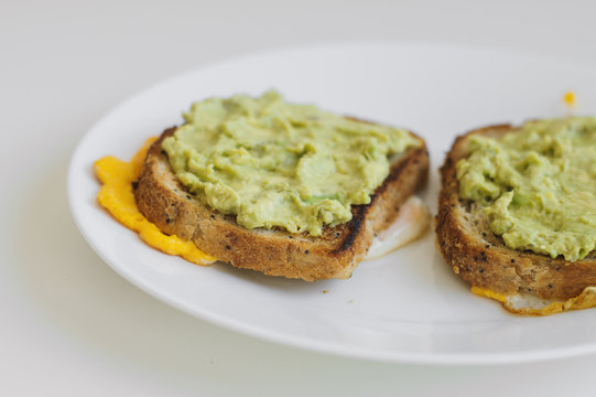 Two toasts with whole grain quinoa bread, smashed avocado and chicken eggs on white plate. Breakfast meal