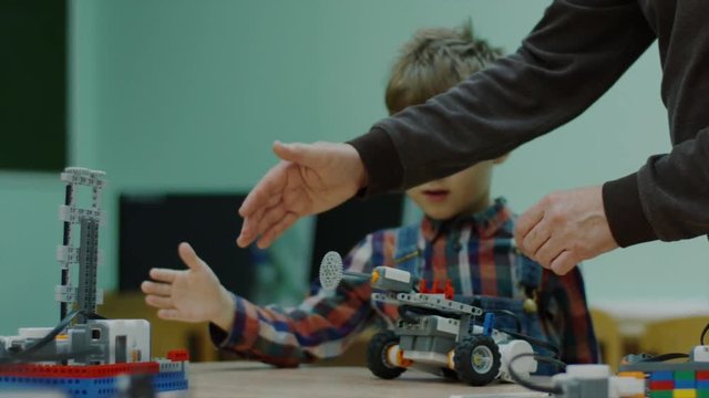 Close up shot of a teacher helping young boy with a robots in school science club. Small blocks car riding on a table. Project for engineering club in school.