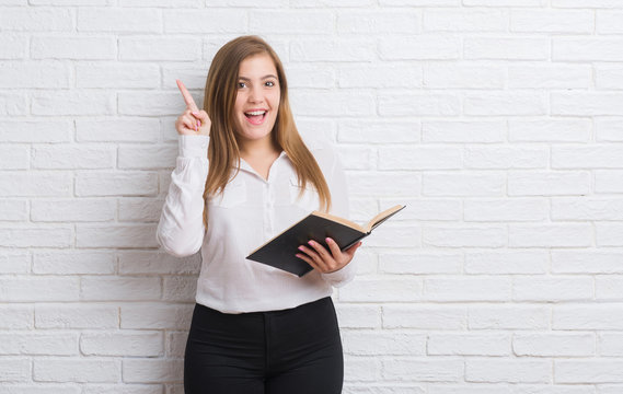 Young adult woman standing over white brick wall reading a book surprised with an idea or question pointing finger with happy face, number one