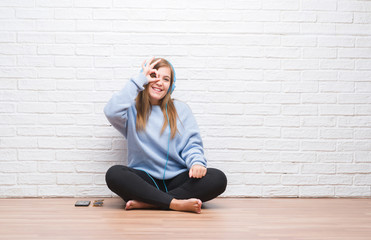 Young adult woman over white brick wall listening music wearing headphones with happy face smiling doing ok sign with hand on eye looking through fingers