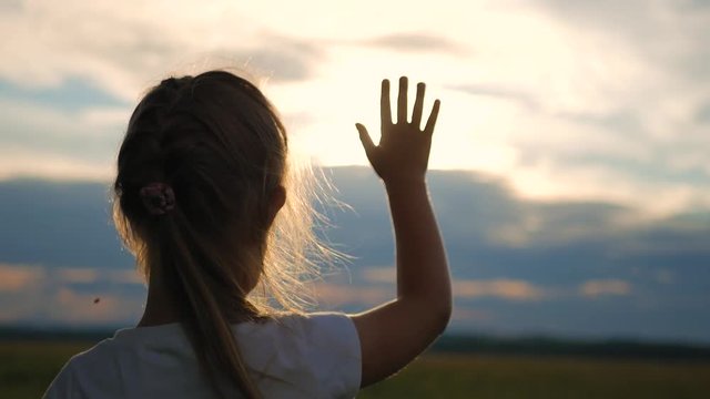 Little blonde girl looks at the sun through her hand. Silhouette of a child at sunset in the field.
