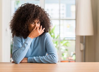 African american woman at home smelling something stinky and disgusting, intolerable smell, holding breath with fingers on nose. Bad smells concept.