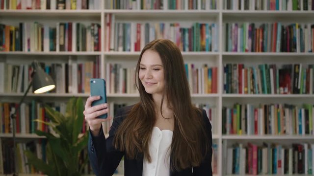portrait of young beautiful woman standing in library using smartphone video chat student waving smiling laughing