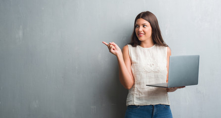Young brunette woman over grunge grey wall using computer laptop very happy pointing with hand and...