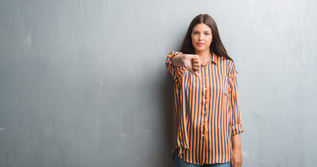 Young brunette woman over grunge grey wall looking unhappy and angry showing rejection and negative with thumbs down gesture. Bad expression.