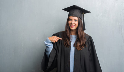 Young brunette woman over grunge grey wall wearing graduate uniform with surprise face pointing...
